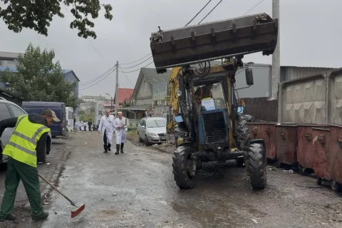 Робітник підмітає мокру вулицю біля трактора і сміттєвих контейнерів