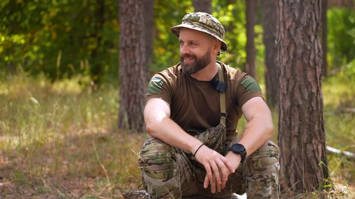 Бородатий військовий у камуфляжі сидить у сосновому лісі й усміхається